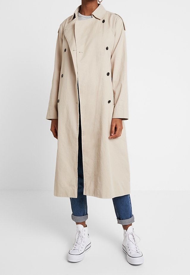 Trench coat beige con colletto, design doppiopetto e bottoni in tessuto. Indossato sopra un jeans e abbinato a sneaker bianche.