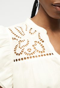 Blouse blanche avec broderie découpée complexe sur l'épaule, gros plan du haut du torse et cheveux tressés.