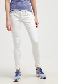 Jeans blancos ajustados con una textura suave, que cuentan con un diseño tradicional de cinco bolsillos y hardware plateado. Combinados con zapatillas deportivas azules.
