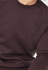 Sudadera de cuello redondo en color marrón con cuello y puños acanalados. Presenta un pequeño detalle de langosta negra bordada en el pecho. Textura suave.