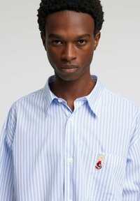 Blå og hvitstripet knappeskjorte med lomme. Har en brodert figur på lommen og en klassisk krage.