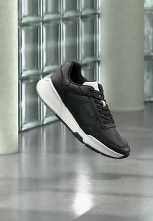 Sneaker in pelle bianca e nera con lacci che fluttua sopra un pavimento in cemento lucido e una parete di blocchi di vetro sullo sfondo.
