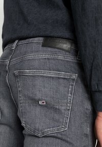 Grå denimjeans har en läderpatch vid midjan och en liten logotyp-patch på bakfickan. Tyget har en texturerad yta.