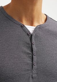 Zbliżenie na osobę w ciemnoszarym podkoszulku henley z czterema częściowo rozpiętymi guzikami, nałożonym na biały podkoszulek.