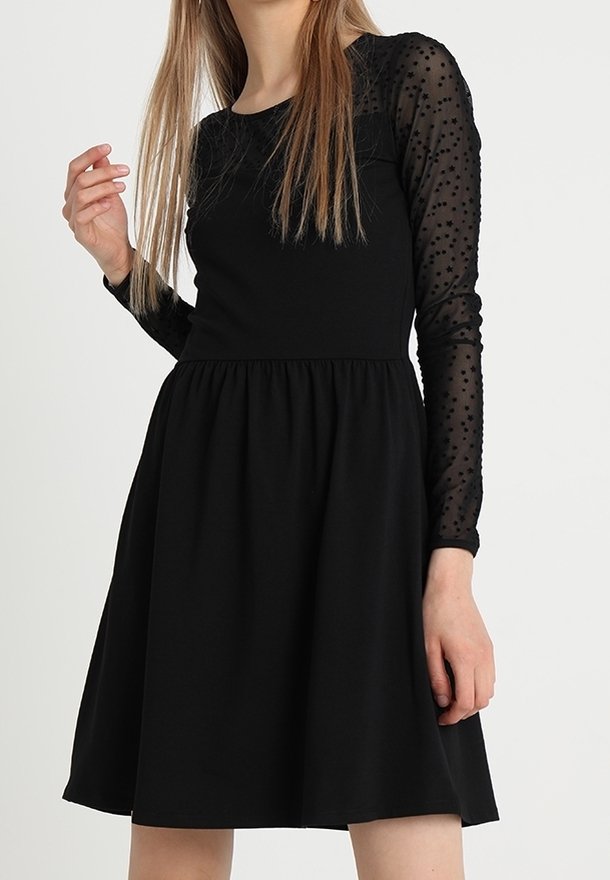 Femme portant une robe noire avec des manches longues transparentes à motifs d'étoiles et une taille froncée, debout devant un fond blanc uni.
