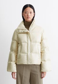 Lys beige dunjakke med høy krage, trykknapper og sidelommer. Har en blank tekstur og kort design.