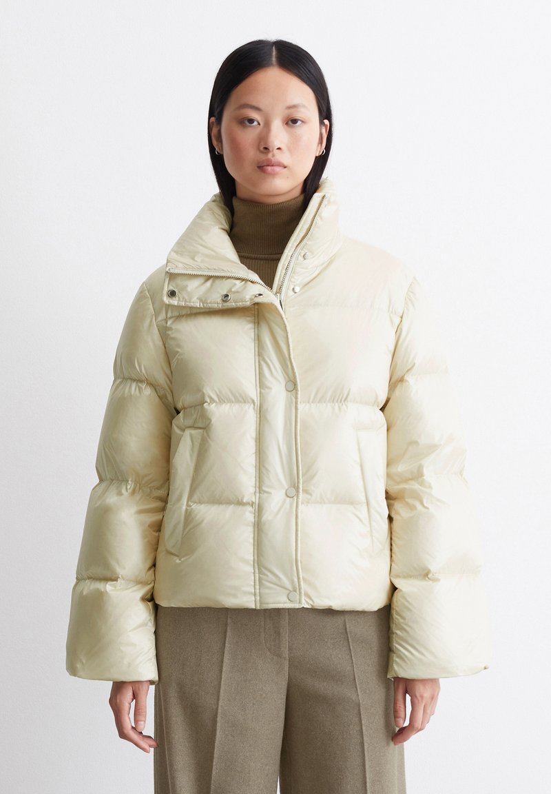 Lys beige dunjakke med høy krage, trykknapper og sidelommer. Har en blank tekstur og kort design.