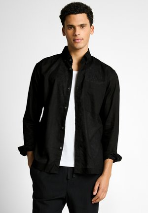 Junger Mann mit lockigem Haar, der ein schwarzes Hemd über einem weißen T-Shirt trägt, steht vor einem schlichten hellen Hintergrund.