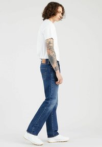 Blauwe spijkerbroeken met een rechte pasvorm, voorzien van een bruine leren patch op de achterkant, gecombineerd met een wit t-shirt met ronde hals en witte sneakers.