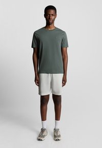 Olijfgroene katoenen t-shirt met korte mouwen, gecombineerd met lichtgrijze shorts. Het model draagt beige sneakers en witte sokken.