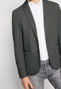 Szary teksturowany blezer z klapą z wycięciem, pojedynczym zapięciem na guzik i dwoma przednimi kieszeniami, noszony na białej koszuli i szarych dżinsach.