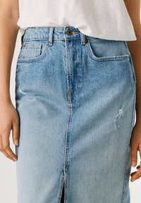 Femeie purtând o fustă din denim albastru deschis cu slit frontal și o cămașă albă băgată înăuntru, mâinile relaxate pe lângă corp.