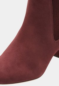 Zbliżenie bordo zamszowego botka do kostki z obcasem typu klocek i elastycznym panelem po bokach.