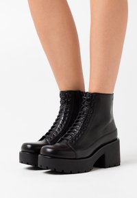 Botas de cuero negras hasta el tobillo con una suela de plataforma gruesa, puntera redonda y cordones en la parte delantera. Presentan ojetes reforzados y una superficie texturizada.