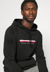 Čierna mikina s kapucňou a šnúrkou, s horizontálnym pruhom v červenej, bielej a modrej farbe nad textom "TOMMY HILFIGER" v bielej farbe. Mäkko tkanina.