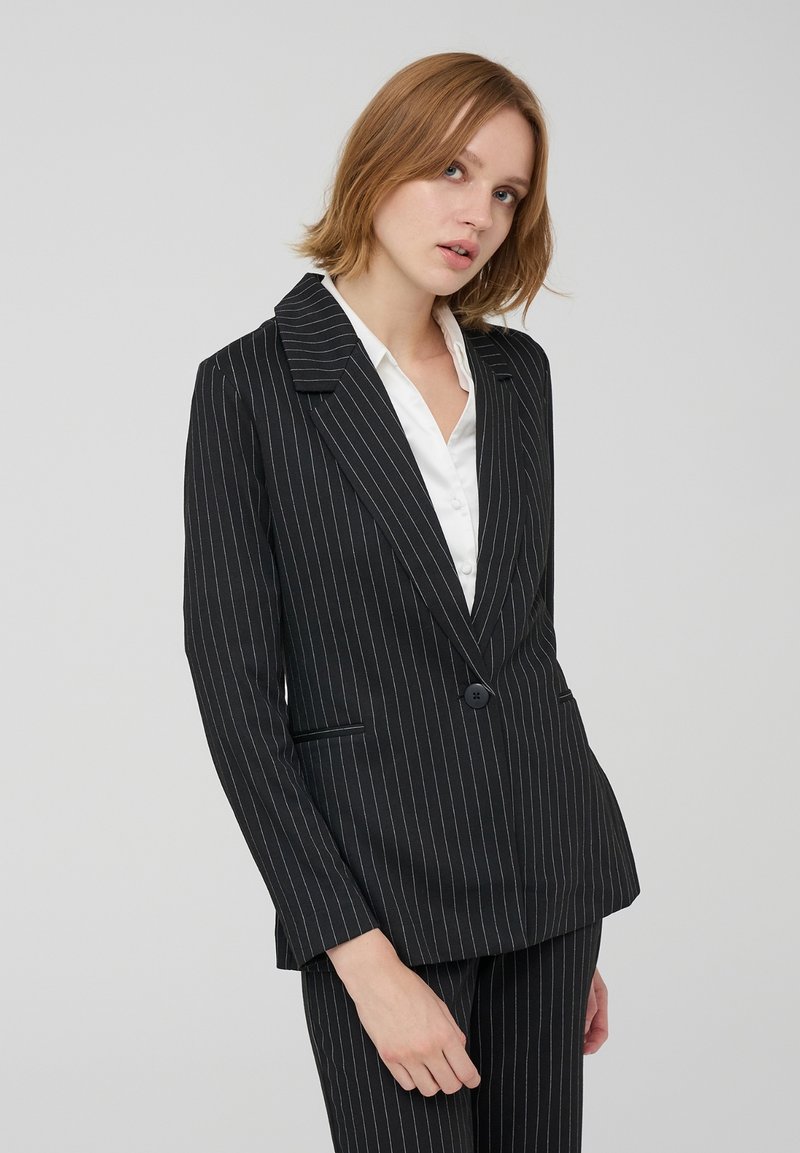 Blazer nero a righe con chiusura a un bottone, revers a punta e due tasche laterali. Realizzato in tessuto strutturato con una finitura liscia.