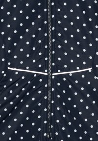 Giacca blu navy con pois bianchi, dotata di zip frontale, accento riflettente e texture del tessuto liscia e leggermente lucida.