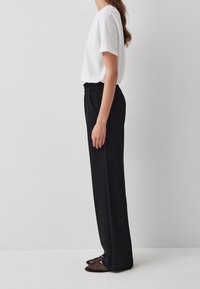Zwarte wijde broek met een elastische tailleband, gedragen met een witte T-shirt met korte mouwen en zwarte mesh ballerina's. Glad materiaal.