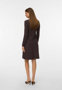 Bruine langemouw jurk met een getailleerde bovenkant en een uitlopende rok, gemaakt van een gladde stof. Schoenen met hakken in een contrasterende donkere kleur.