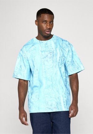 Mand med kort hår iført lys blå, mønstret, oversized t-shirt og mørkeblå jeans mod en ensfarvet baggrund.