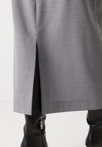 Jupe grise avec une fente arrière laissant voir des bottes en cuir noires avec des fermetures éclair portées en dessous sur fond blanc.