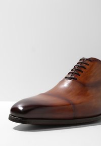 Chaussure en cuir marron à lacets avec une finition lisse, bout pointu et bords arrondis. Présente un dégradé subtil du marron foncé au marron clair.