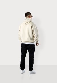 Sweat à capuche beige avec cordon de serrage, coupe décontractée et poignets côtelés, porté par-dessus une chemise blanche. Associé à un pantalon noir et des baskets blanches.