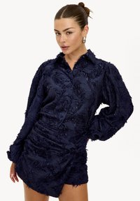 Robe-chemise texturée bleu marine avec col, manches longues bouffantes et détails complexes sur le tissu, dotée d'une taille froncée.