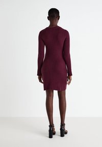 Bordový žebrovaný pletený šat s dlouhými rukávy, přiléhavým střihem a kulatým výstřihem, končící nad koleny. Nosený s černými sandály na vysokém podpatku.