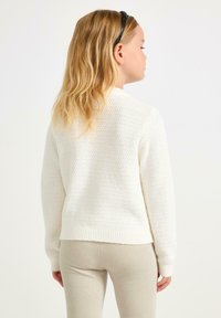 Kind met lang blond haar, gekleed in een witte textuurtrui en beige broek, van achteren gezien tegen een effen lichte achtergrond.