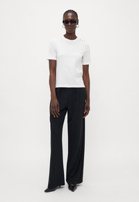 White short-sleeve t-shirt with round neckline and three decorative buttons on the left shoulder, paired with loose black trousers and heels.
