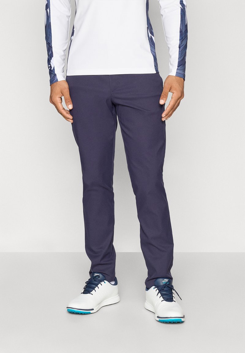 Men's navy pants made from a smooth fabric. They feature a fitted design with a slight stretch, paired with white athletic shoes.