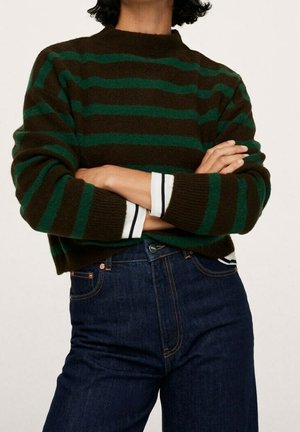 Person stående med armene over kors iført en mørkebrun og grøn stribet sweater over en hvid skjorte med sorte striber og mørkeblå jeans.