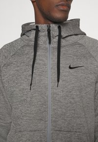 Hombre vistiendo una sudadera con cremallera gris de Nike con cordones negros, mostrando la parte superior del torso y parte de la cara.