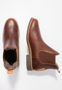 Botas de cuero marrón hasta el tobillo con una suela marrón texturizada, paneles laterales elásticos y un forro suave. Presentan una tira de agarre en el talón y detalles de costura.