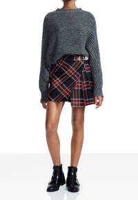 Grijze geribbelde sweater met een losse pasvorm, gecombineerd met een zwart-rode geruite wikkelrok met plooien en een zilveren gesp riem. Zwarte enkelhoge laarzen.