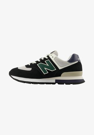 Svart och grå New Balance-sneaker med grön "N"-logotyp, vita snören och vadderad sula, sedd från sidan mot en vit bakgrund.
