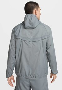 Giacca grigia leggera con cappuccio, vestibilità ampia, polsini e orlo elastici, caratterizzata da una texture liscia e dettagli di design minimalisti sul retro.