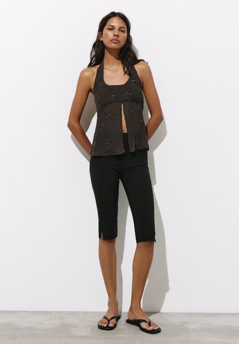 Femme debout contre un mur blanc portant un haut sans manches noir à col halter avec des motifs d’araignées et un pantalon noir ajusté aux genoux avec des fentes.