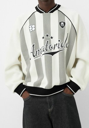 Person trägt einen weiß-schwarz gestreiften Pullover mit dem Schriftzug "Amatoriale" und Sternsymbolen, kombiniert mit dunklen Jeans, die Hände in den Taschen.