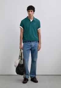 Polo verde con mangas enrolladas, jeans de mezclilla azul claro, zapatos marrones y una bolsa de mano con múltiples bolsillos. Diseño simple y estilo casual.