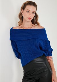 Modra rebrasta off-shoulder pulover iz mehkega pletenega materiala, z obsežnimi rokavi in strukturiranim ovratnikom, v kombinaciji z črnimi usnjenimi hlačami.