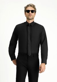 Camisa negra de manga larga, con cuello abotonado y un solo bolsillo en el pecho. Combinada con una corbata negra texturizada y gafas de sol.