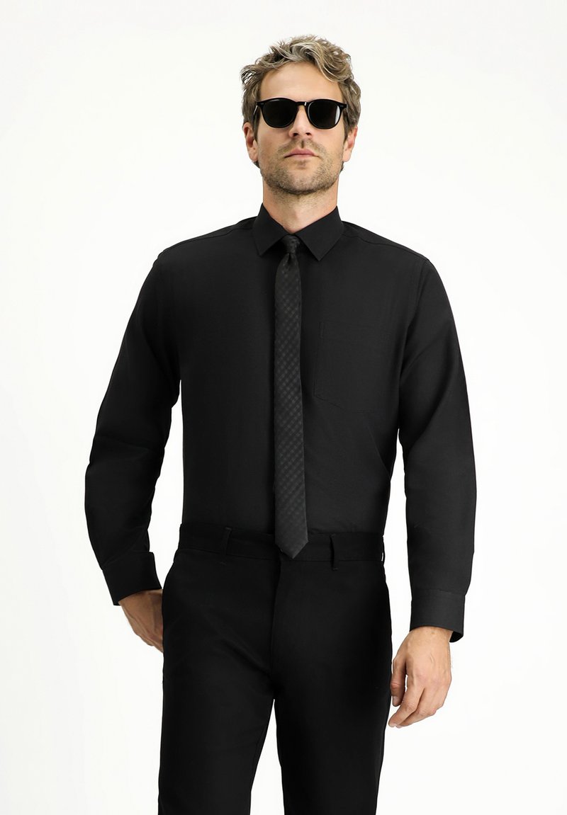Camisa negra de manga larga, con cuello abotonado y un solo bolsillo en el pecho. Combinada con una corbata negra texturizada y gafas de sol.