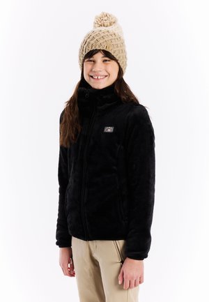 Zwarte fleece jas met een hoge kraag en rits, met een klein logo patch. Gekleed met een beige gebreide beanie met een pompon bovenop.