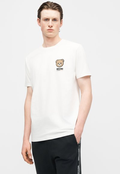 Νέος άνδρας που φοράει λευκό t-shirt Moschino με μικρό λογότυπο αρκούδας στο στήθος και μαύρο παντελόνι με πλαϊνή επωνυμία, στέκεται με το ένα χέρι στην τσέπη.