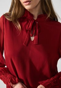Rød bluse med høy hals, med knyttet båndlukking og teksturert stoff. Lange ermer med volanger ved ermene gir ekstra detaljer.