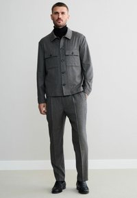 Grau melierte Wolljacke mit zwei Brusttaschen, schwarzen Knöpfen, kombiniert mit passenden Tailor-Hosen und schwarzen Lederschuhen.