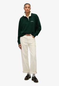 Zielona koszula polo z długim rękawem i kontrastowym kremowym kołnierzykiem, zestawiona z kremowymi spodniami typu wide-leg i ciemnymi butami. Zawiera haftowany tekst.