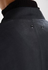 Abrigo negro con una tela texturizada, que presenta un alto cuello y dos broches de metal en la parte posterior para un detalle adicional.
